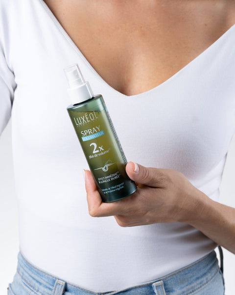 Žmogus, laikantis „Luxe Oil“ purškiamos dulksnos buteliuką baltame fone