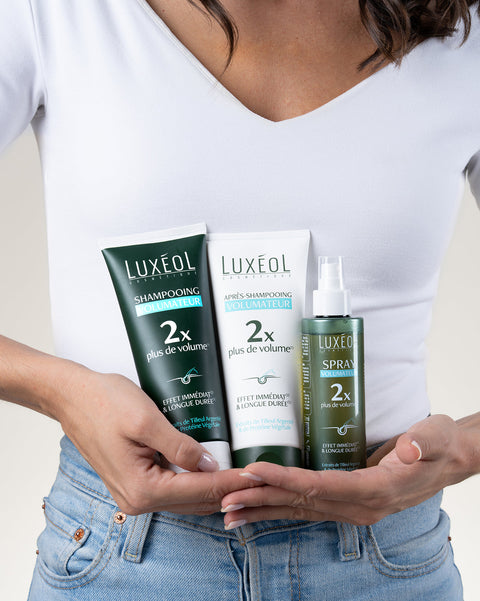 Asmuo, laikantis Luxéol plaukų priežiūros produktus, įskaitant šampūną, kondicionierių ir purškiamą dulksną.